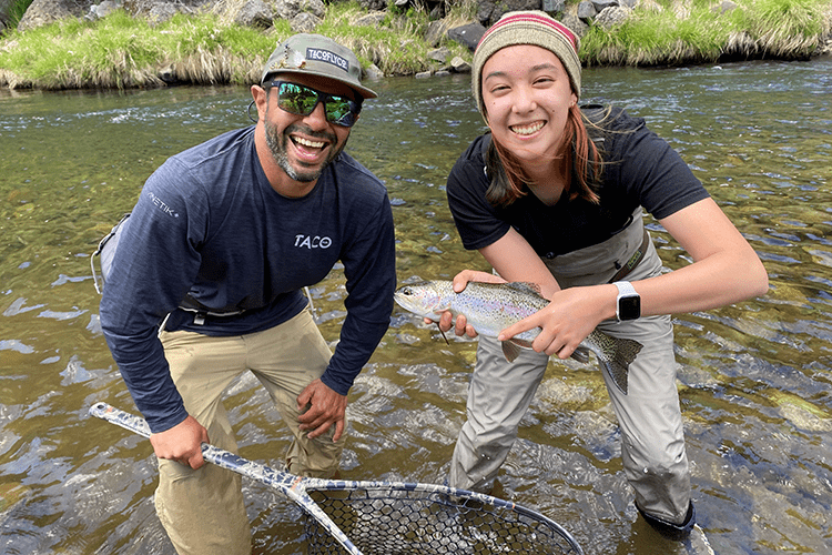 2 people with smiling faces standing in river with rainbow trout