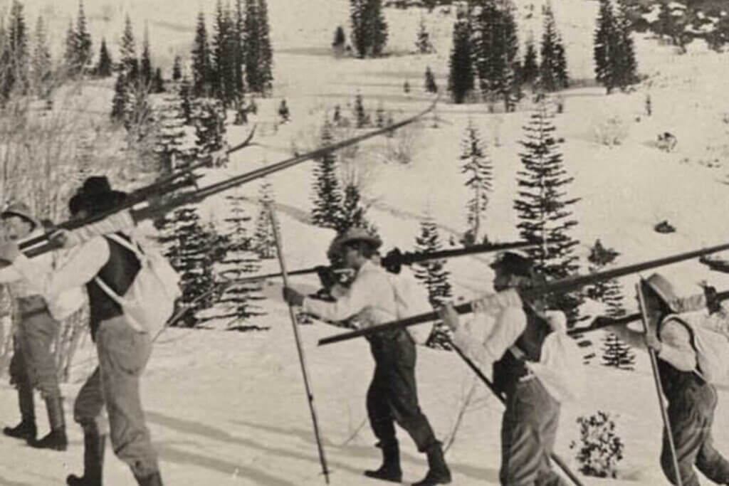 Historical photo of longboard skiers carrying skis and climbing mountain